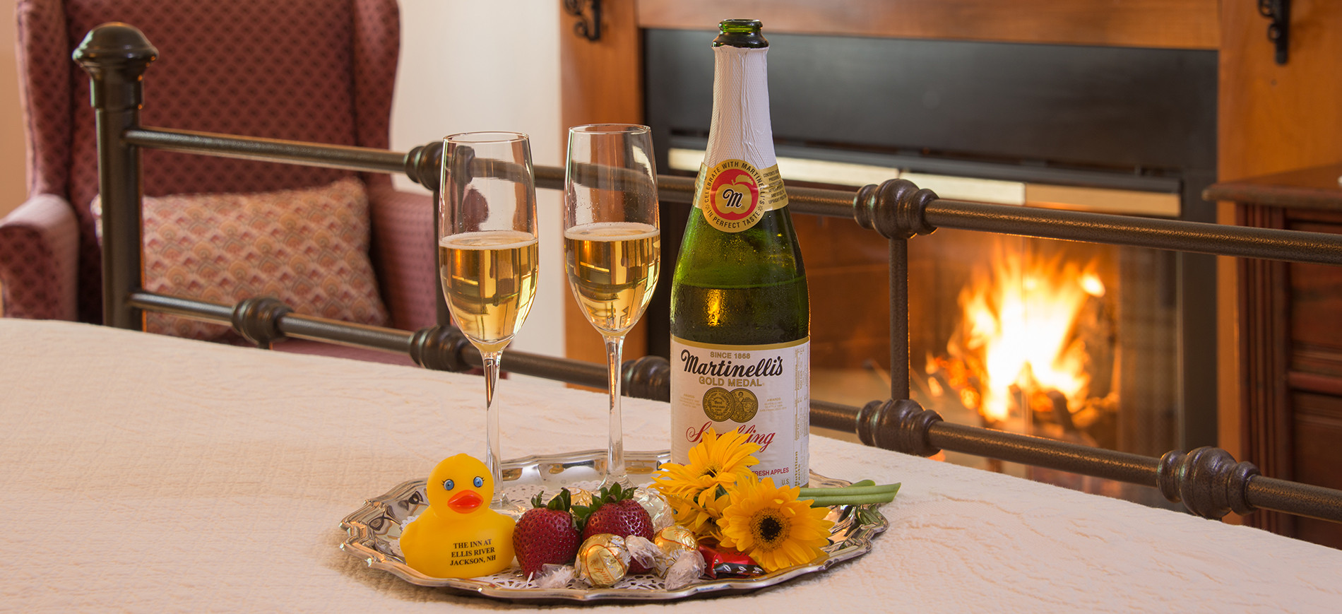 Glasses & bottle of cider on silver tray with rubber duck, strawberries, chocolates. Tray on bronze bed with fireplace behind