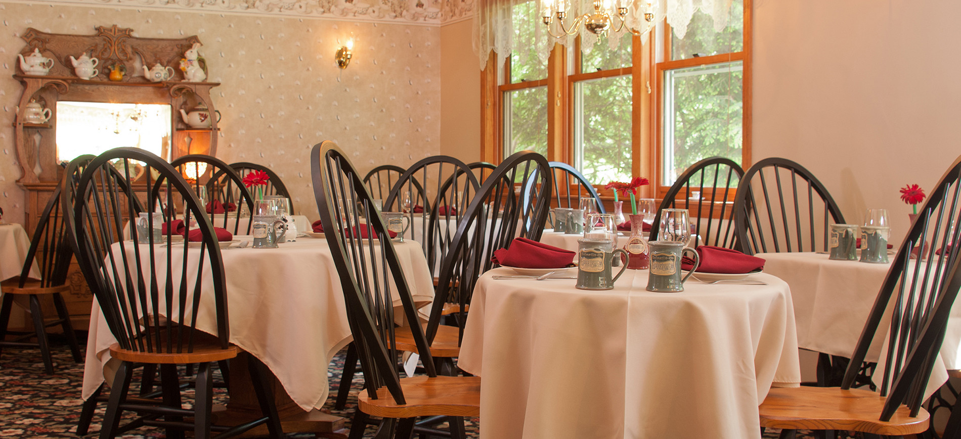 Breakfast room with ivory table cloths, burgundy napkins, sage green mugs, black & oak Windsor chairs, hutch with teapots in background