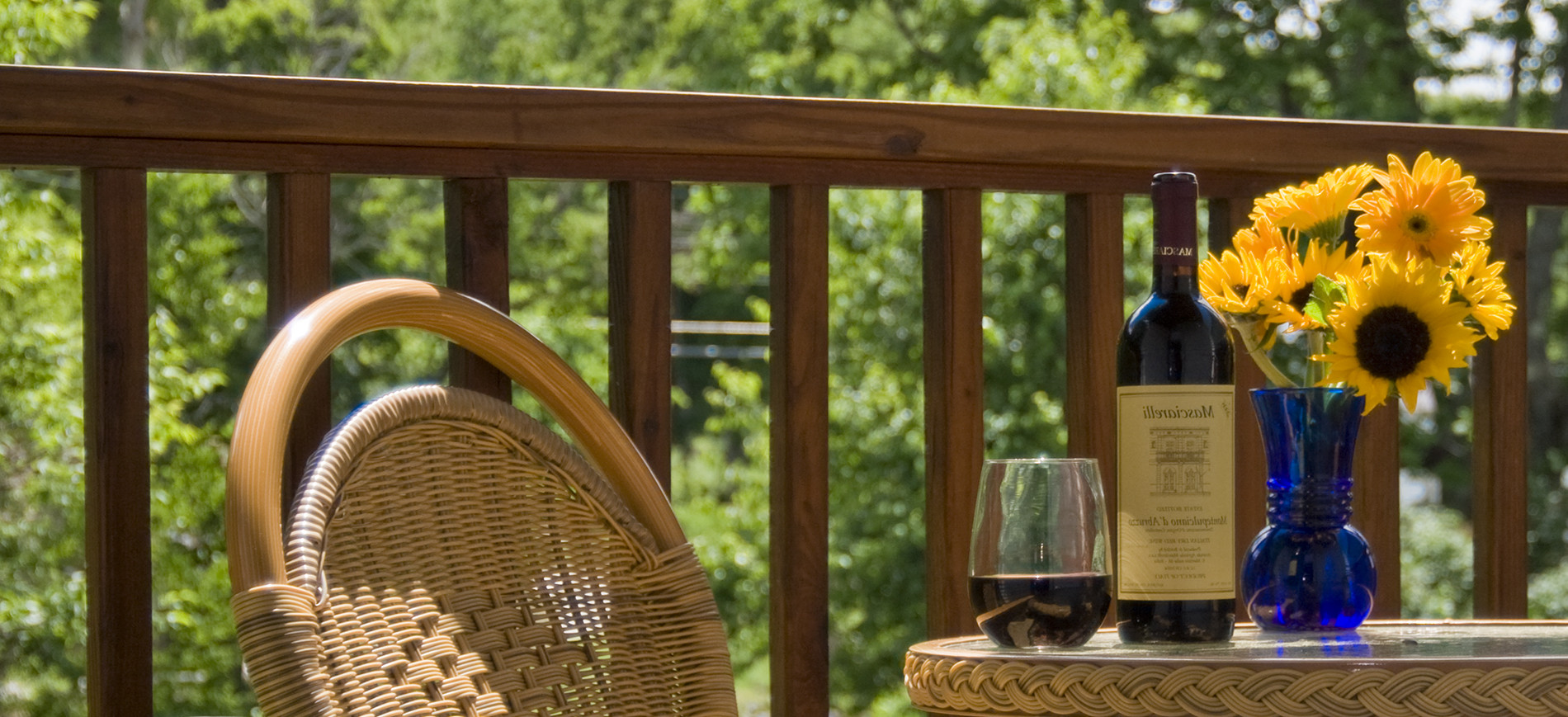 wicker chair, table top with glass of red wine, bottle, yellow flowers in blue vase, balcony railing & trees in background