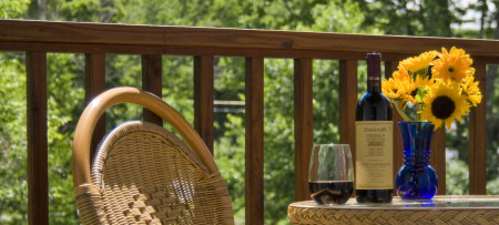 wicker chair, table top with glass of red wine, bottle, yellow flowers in blue vase, balcony railing & trees in background