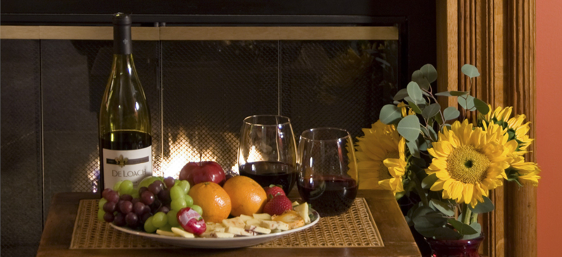 wine bottle, fruit & cheese plate, two glasses wine, sunflowers with fireplace behind