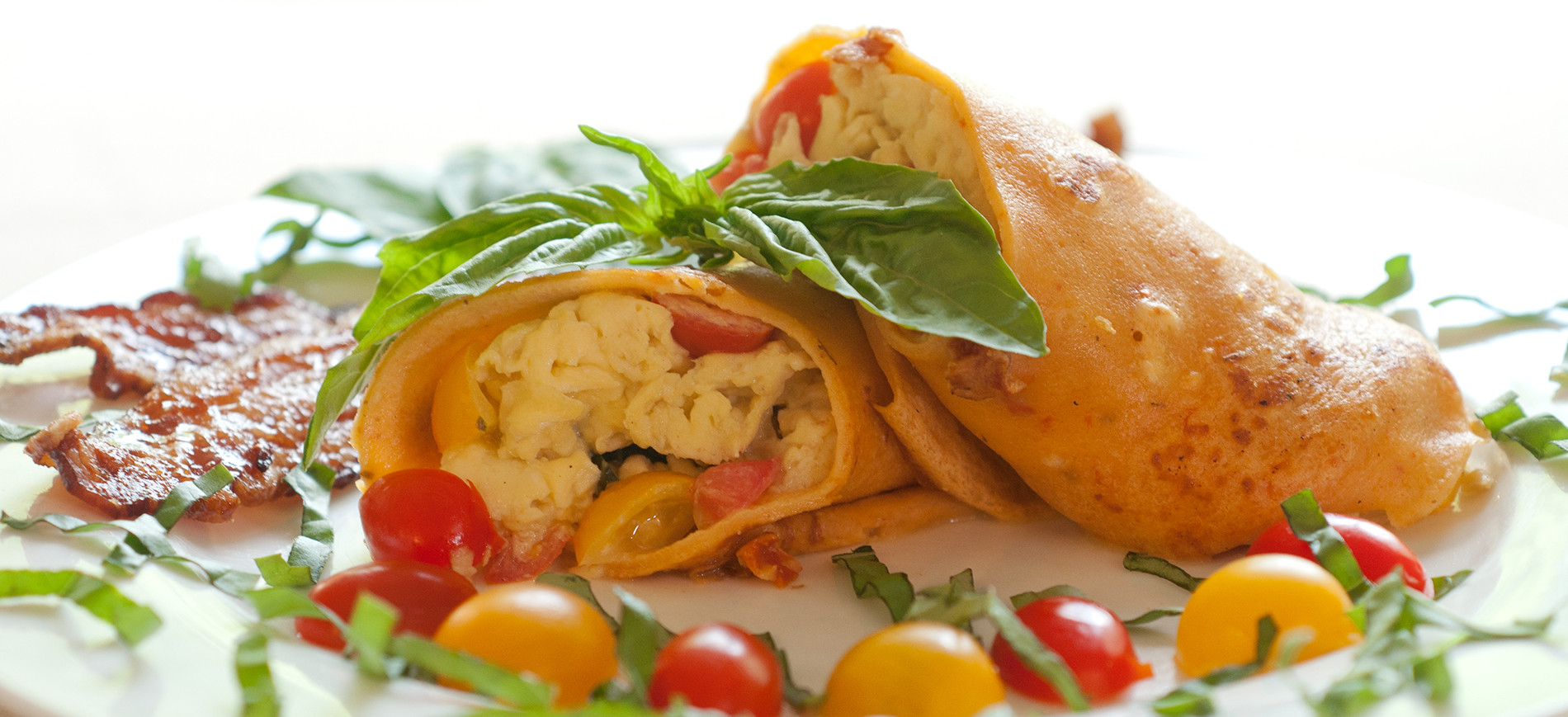 Two halves Crepe Caprese with basil garnish stacked on white plate with ring of red & yellow cherry tomatoes and basil chiffonade