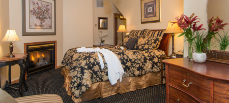 Chaise, table with lamp, fireplace with picture above, queen sleigh bed with gold & black bedding, chest with mirror above