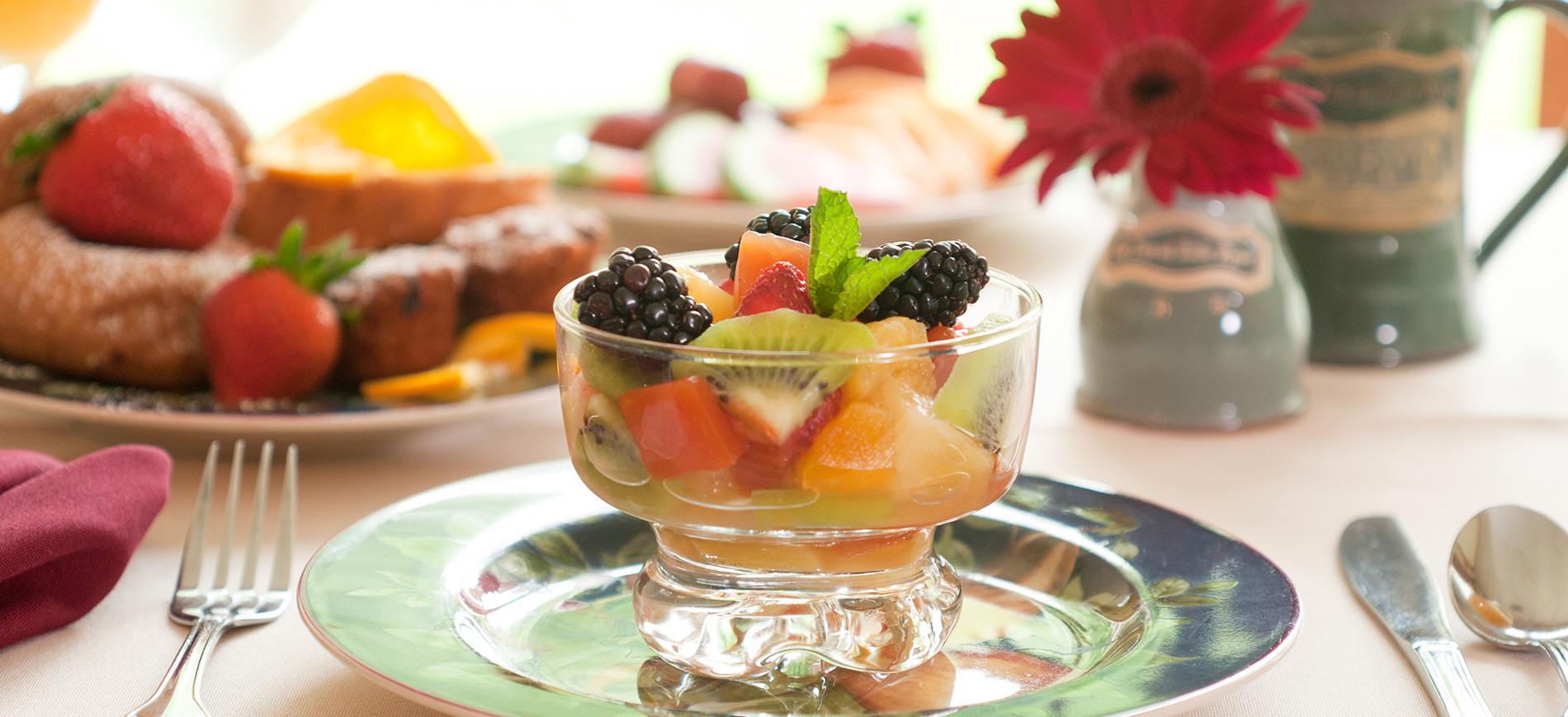 fruit cup on plate, vase with red flower and mug behind right, bread and strawberries behind left