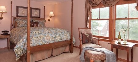 queen four poster bed with aqua & beige bedding, wall lamps on each side of bed, beige arm chair with ottoman, aqua shawl, table with wine & flowers