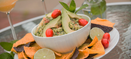 Guacamole with cherry tomato garnish in white bowl surrounded by yellow and blue corn chips