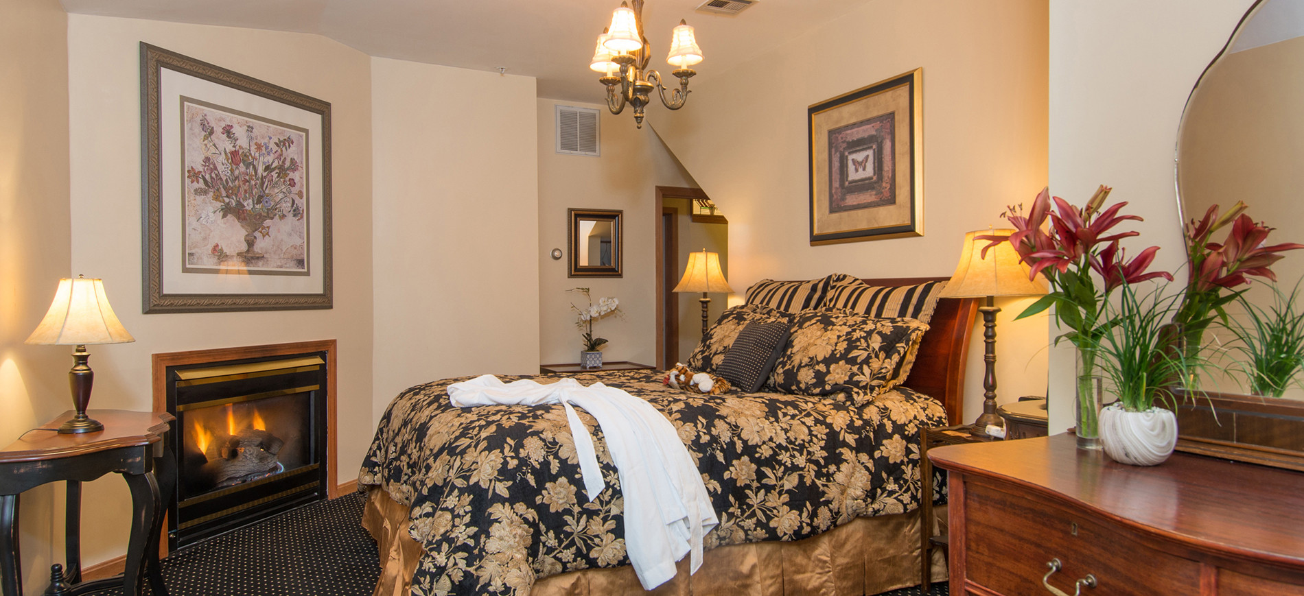 Fireplace with picture above, queen bed with gold & black bedding & white robe below chandelier