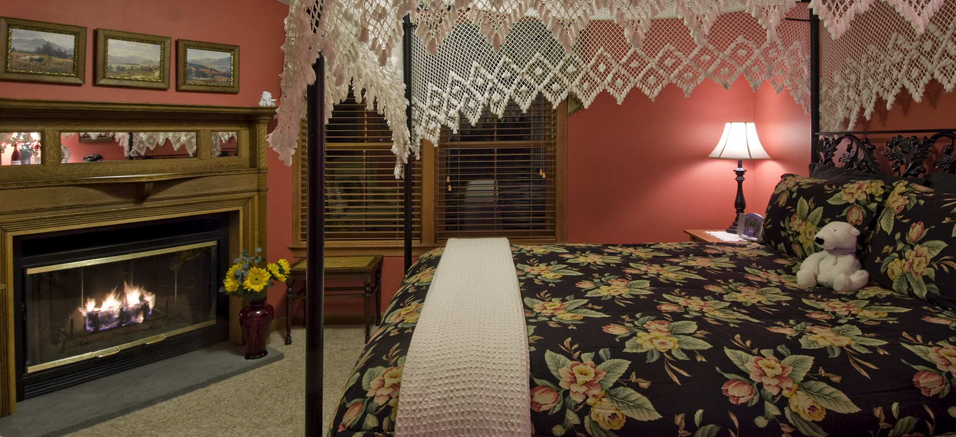 Guest room with salmon walls, fireplace with antique mantel & pictures above, black four poster bed with white lace canopy & black, salmon & green bedding