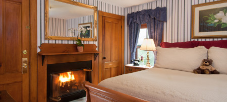 Guest room with blue & white striped wallpaper, fireplace with mirror above, closet door, window with blue drapes, night stand with brass lamp, king bed with ivory bedding and picture above