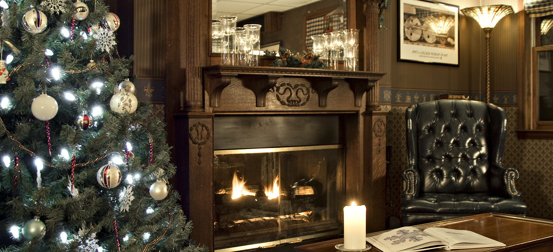 Xmas tree on left, fireplace with mantel, wing chair, coffee table with candle and open book