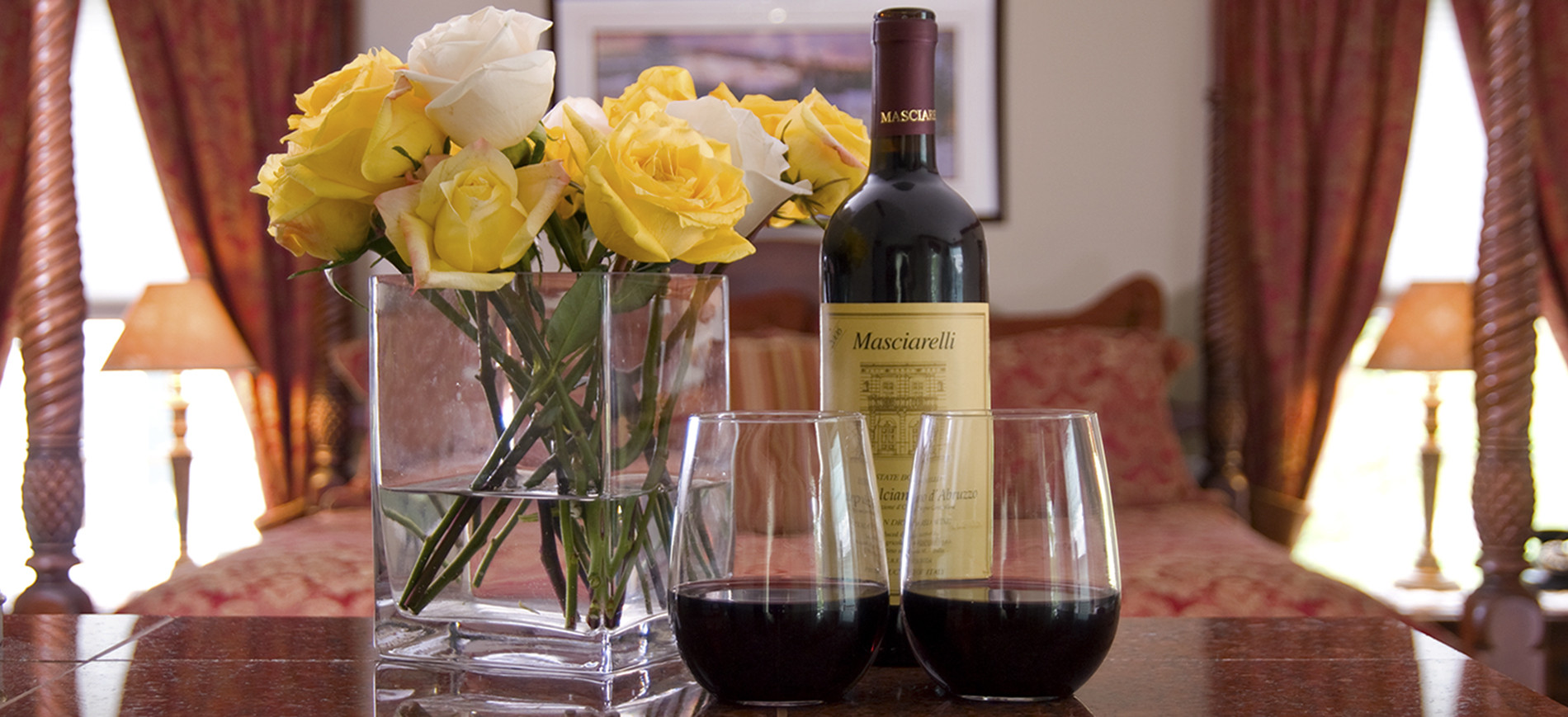 White & yellow roses in clear vase on left, bottle of wine & two glasses of red wine on right