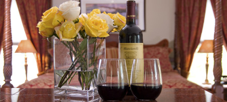 White & yellow roses in clear vase on left, bottle of wine & two glasses of red wine on right