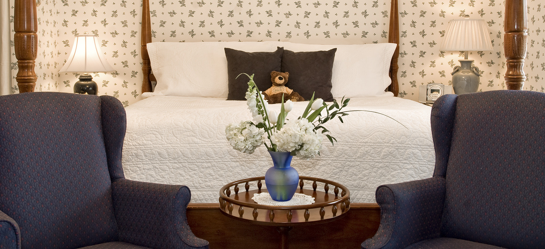 Guest room with white & blue wallpaper, king bed with white bedding in back of blue wing chairs with round table with flowers