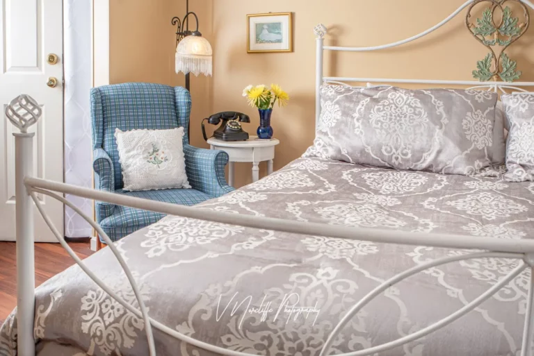 bedroom showing a white metal bed frame with a patterned duvet, a blue armchair, and a nightstand with a lamp.