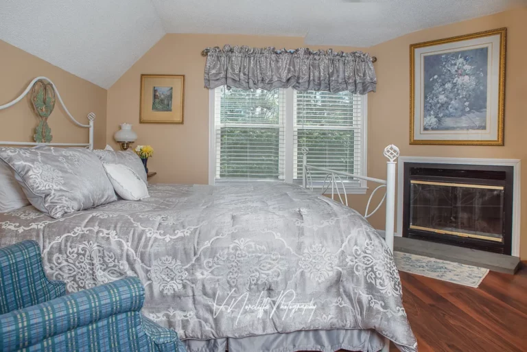 bedroom with a patterned bedspread, blue armchair, window with shutters, and a fireplace.