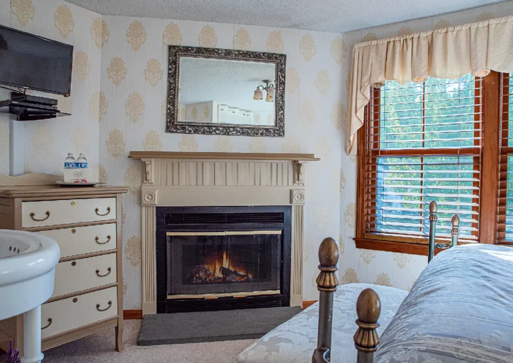 Bedroom with a fireplace, TV, and window looking out to greenery.