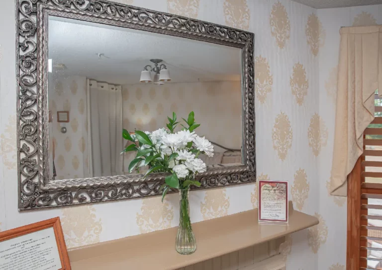 Dressing table with an ornate mirror, a vase of white flowers, and patterned wallpaper.